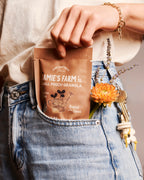 A person wearing blue jeans and a cream top holds a brown pouch of Jamie's Farm Small Pouch Granola in their pocket, with keys and dried flowers hanging from the pocket.